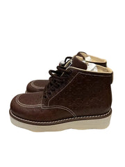A pair of dark brown Louis Vuitton monogram embossed leather ankle boots with white contrast stitching and off-white platform soles, displayed on a white textured surface against a gray wall.
