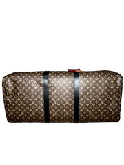 A Louis Vuitton monogram canvas duffel bag with black leather straps, resting on a wooden surface against a gray textured background.