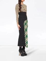 A model wearing a long-sleeve knit maxi skirt with a black body and green tartan plaid side panels, paired with a beige and brown plaid top, against a plain white studio background.
