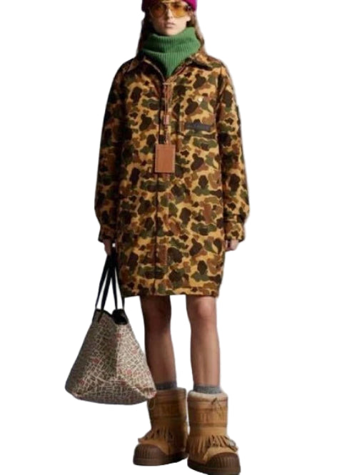 Person wearing a camouflage dress with a green turtleneck, holding a patterned bag, and wearing brown boots on a gray background.