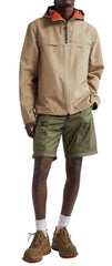 A man wearing a beige hooded jacket, olive green shorts, and brown boots, shown from the waist up against a plain light background.