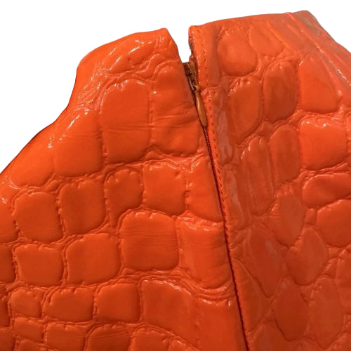 Close-up of a bright orange crocodile-embossed leather item being held by a hand, showing the glossy textured surface and a gold-toned zipper.