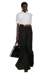 A model wearing a white short-sleeve button-up shirt, black wide-leg trousers, and a gold belt, holding a black handbag against a light-colored wooden background.