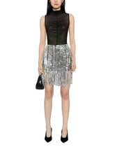 A model wearing a sleeveless black sheer mock-neck top paired with a silver sequined fringe mini skirt, holding a small black handbag against a plain white background.