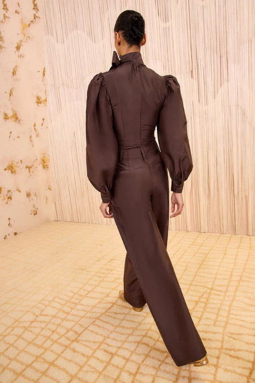 A model viewed from behind wearing a dark brown satin-like jumpsuit with dramatic puffed sleeves and wide-leg trousers, standing in a cream-colored room with textured walls and a patterned rug.