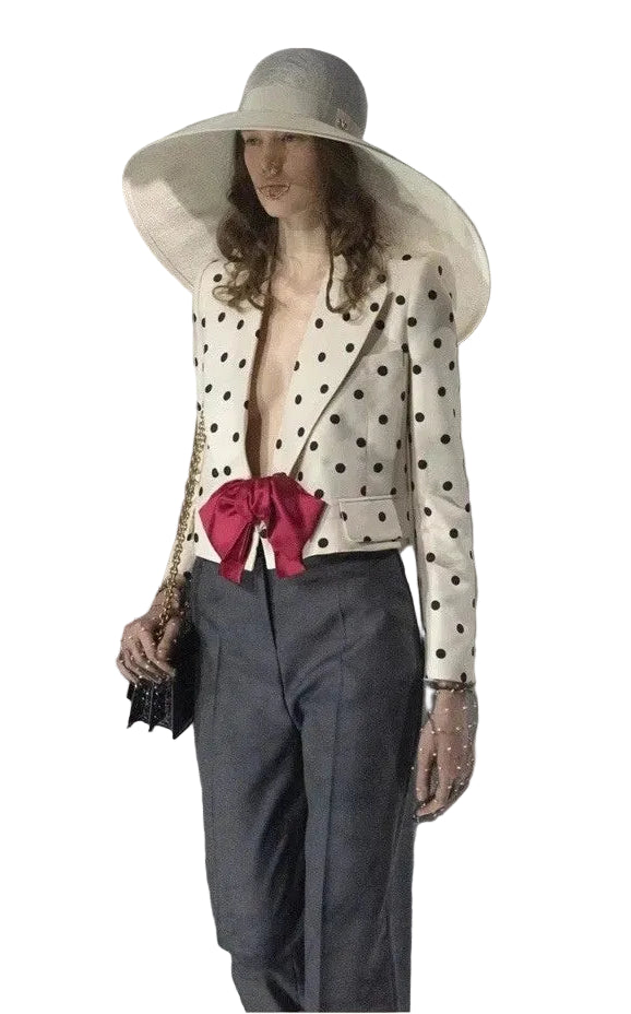 A model wearing a wide-brimmed white sun hat, a cream blazer with black polka dots and a large red bow at the waist, dark gray trousers, and holding a black clutch bag.