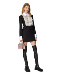 Woman wearing a black dress with white ruffled collar, black knee-high boots, and holding a pink handbag on a white background.