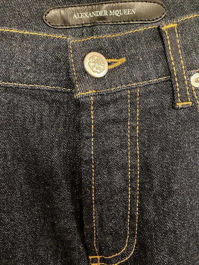 Close-up of dark indigo denim jeans showing the waistband, button, yellow contrast stitching, and an Alexander McQueen leather brand tab.