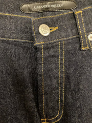 Close-up of dark indigo denim jeans showing the waistband, button, yellow contrast stitching, and an Alexander McQueen leather brand tab.