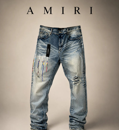 Light blue distressed denim jeans by Amiri, featuring colorful thread details on the left leg, ripped details on the right thigh, and a black Amiri brand tag hanging from the waistband, set against a neutral beige background with the Amiri logo at the top.