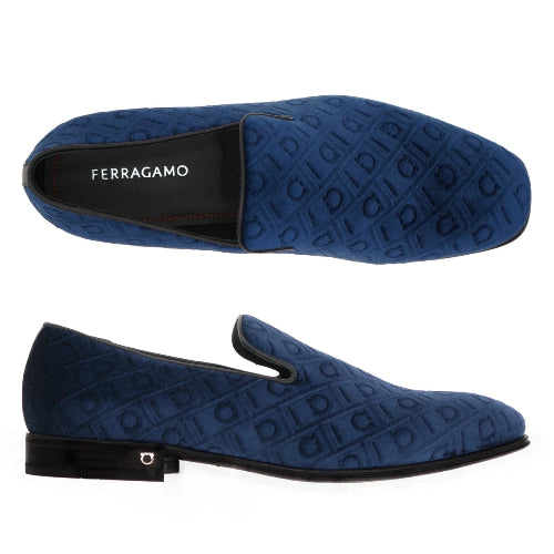 A pair of blue suede slip-on loafers shown from the side and top views against a black background. The brand name 'FERRAGAMO' is visible on the black heel tab.