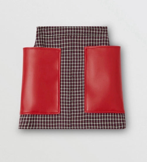 A structured mini skirt featuring a dark brown and white micro-check patterned body with two contrasting bright red faux-leather front pockets, shown against a light gray background.