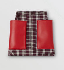 A structured mini skirt featuring a dark brown and white micro-check patterned body with two contrasting bright red faux-leather front pockets, shown against a light gray background.