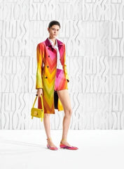 A model stands against a white textured wall wearing a vibrant, multicolored geometric print trench coat and matching shorts, holding a small yellow handbag.