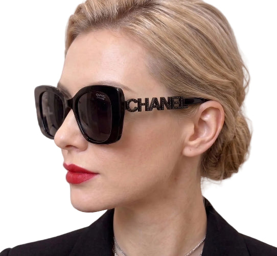 A close-up of a woman wearing oversized black sunglasses with a prominent CHANEL logo on the frame, set against a dark background.