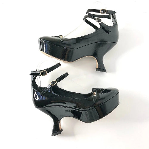 A pair of glossy black patent leather Mary Jane platform heels with ankle straps and buckles, displayed on clear plastic mannequin legs against a white background.