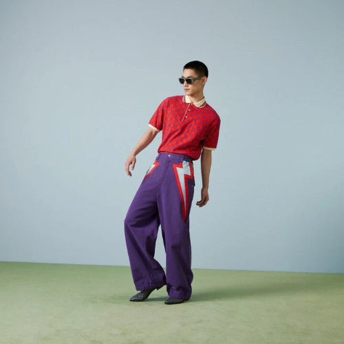 A model wearing a red short-sleeve knit polo shirt and purple wide-leg trousers with red and white graphic details, standing against a light gray studio backdrop with a green floor.