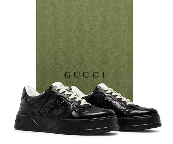 Black Gucci low-top sneakers with white laces, displayed in front of a green Gucci box on a white background.