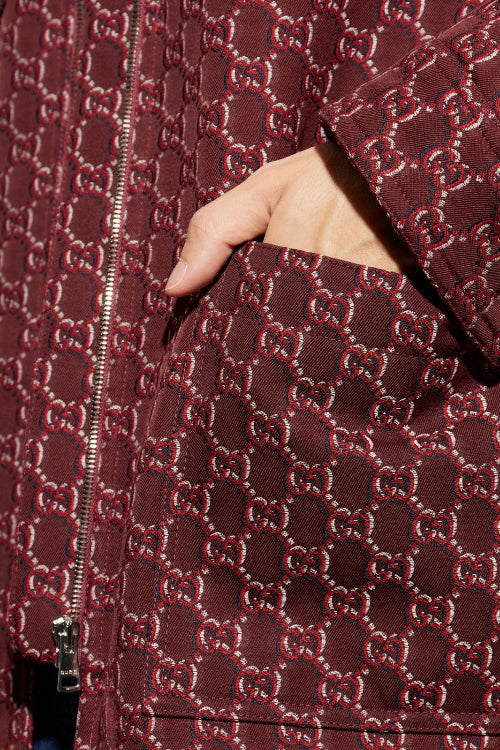 Close-up of a dark brown quilted jacket featuring an allover red and beige Gucci GG monogram pattern, with a hand tucked into a pocket.