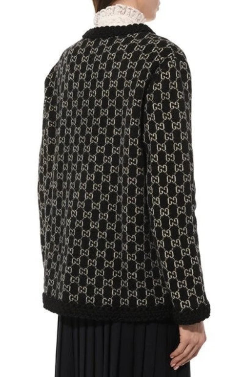 A person shown from the back wearing a black knit sweater with an allover beige interlocking monogram pattern and a black lace-trim collar, paired with a pleated black skirt against a plain white background.