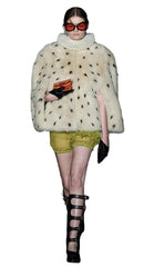 Model wearing a white fur coat with black spots, green shorts, and black high-heeled shoes on a runway.