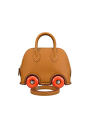 A structured tan leather handbag designed to look like a small car, featuring bright orange wheels with silver hubcaps and a matching tan leather shoulder strap resting in front, set against a neutral gray background.