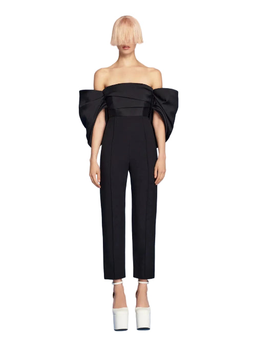 Black off-shoulder jumpsuit with a large bow detail on a blue background