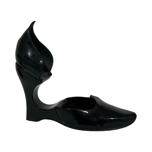A single black patent leather wedge heel shoe with a unique sculptural, flame-like heel and open side cutout, shown against a plain white background.
