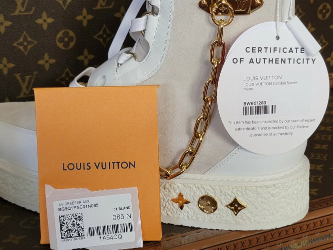 A Louis Vuitton white leather high-top sneaker shown with its orange Louis Vuitton box, authenticity certificate tag, and gold chain detail against a brown monogram background.