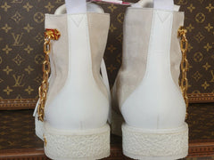 A pair of white leather and suede ankle boots with chunky white platform soles and gold chain details, shown from the back against a brown Louis Vuitton monogram background.
