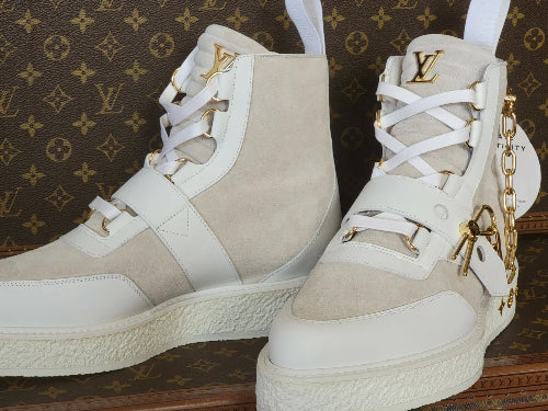 A pair of high-top platform sneakers in beige and white leather with gold-tone hardware, including an LV logo on the tongue and a decorative gold chain detail, set against a brown Louis Vuitton monogram background.