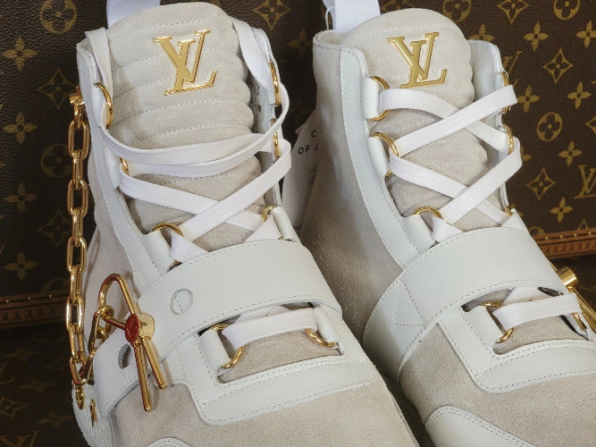 A pair of Louis Vuitton high-top sneakers in off-white leather and suede with gold-tone hardware, featuring the gold LV logo on the tongue and a gold chain detail on the side, set against a brown monogram background.