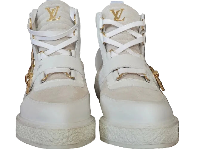 A pair of white Louis Vuitton high-top sneakers with gold-tone hardware and the iconic LV logo on the tongue, set against a brown monogram-patterned background.