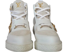 A pair of white Louis Vuitton high-top sneakers with gold-tone hardware and the iconic LV logo on the tongue, set against a brown monogram-patterned background.