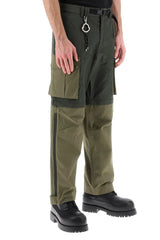 A person wearing olive green and black cargo-style pants with black work boots, shown from the waist down against a light gray studio background.