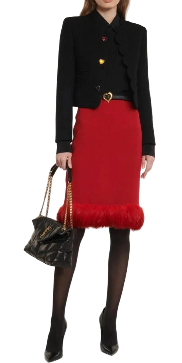 A model wearing a black cropped jacket with gold heart buttons, a red pencil skirt with a red faux-fur hem, black tights, and holding a black quilted handbag with a gold chain strap.