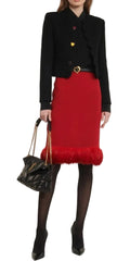 A model wearing a black cropped jacket with gold heart buttons, a red pencil skirt with a red faux-fur hem, black tights, and holding a black quilted handbag with a gold chain strap.