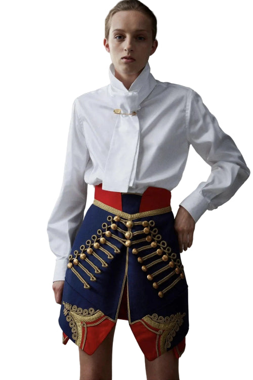 A model wearing a white long-sleeve high-collar shirt tucked into a highly decorative navy blue mini skirt with red panels and gold military-style embroidery and epaulets, set against a plain gray studio background.