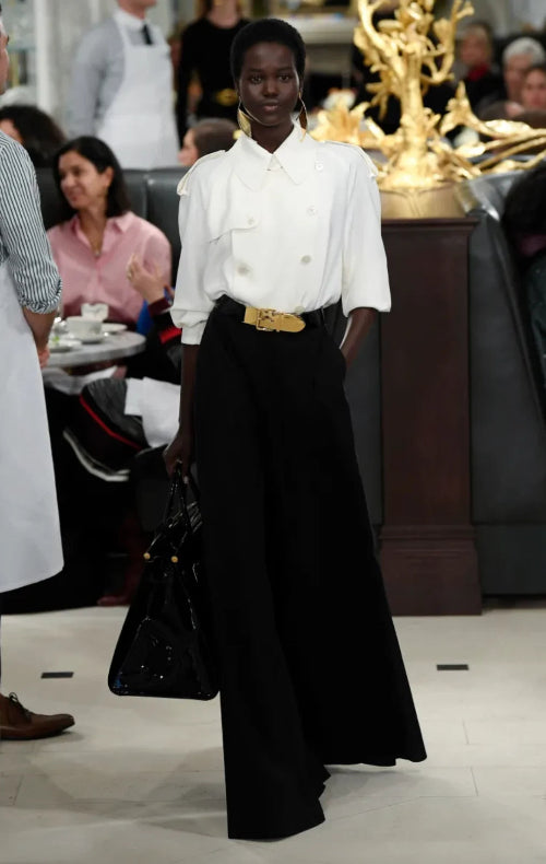 A model on a runway wearing a white long-sleeve top and wide-leg black trousers, photographed in a fashion show setting with an audience in the background.