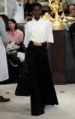 A model on a runway wearing a white long-sleeve top and wide-leg black trousers, photographed in a fashion show setting with an audience in the background.