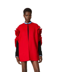 A model wearing a vibrant red mini dress with dramatic ruffled shoulder details and contrasting black sleeves, set against a plain off-white background.