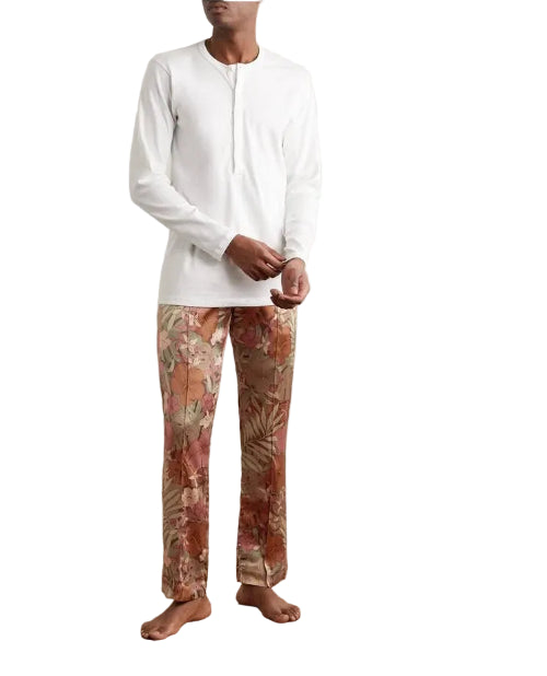 A man wearing a white long-sleeve henley shirt and patterned trousers with a tropical floral print in shades of brown, beige, and orange, standing barefoot on a wooden parquet floor against a plain off-white wall.