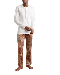 A man wearing a white long-sleeve henley shirt and patterned trousers with a tropical floral print in shades of brown, beige, and orange, standing barefoot on a wooden parquet floor against a plain off-white wall.