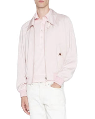 A model wearing a light pink zip-up jacket over a matching pink polo shirt and white trousers, photographed against a plain light gray studio background.