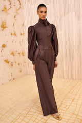 A model wearing a dark brown high-neck jumpsuit with dramatic puffed sleeves and wide-leg trousers, standing in a luxury interior with a beige patterned floor and textured wall.