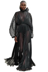 Model wearing a black, flowing dress on a fashion runway with an audience.