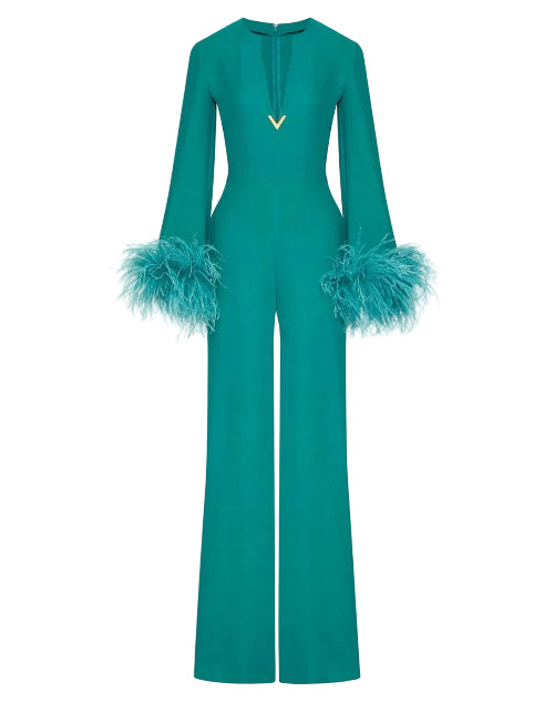 A teal green long-sleeve jumpsuit with a split neckline, a small gold V-shaped metal accent at the center chest, and voluminous feathered cuffs, shown against a solid light beige background.