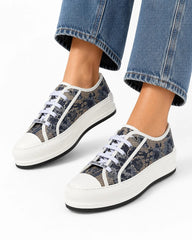 A pair of low-top canvas sneakers with a dark gray and black abstract pattern, white rubber soles, and white laces, shown on a model's feet with blue jeans cropped at the top.