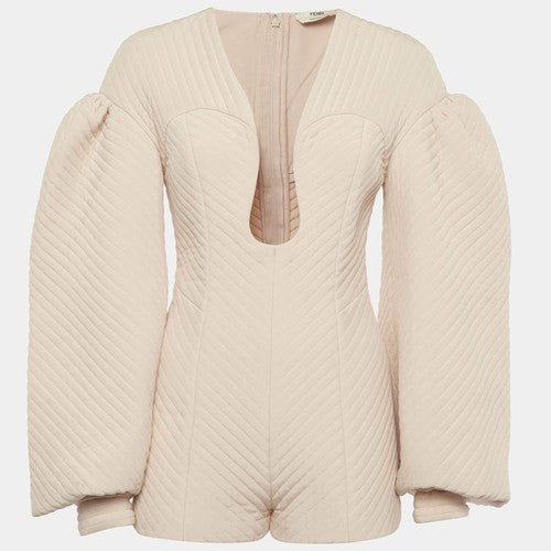 A cream-colored quilted jumpsuit with long puffed sleeves, a deep V-neckline, and a front zipper, shown against a light gray background.