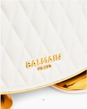 Close-up of a white quilted Balmain Paris handbag with gold lettering and a gold metallic trim, showing the brand logo clearly.